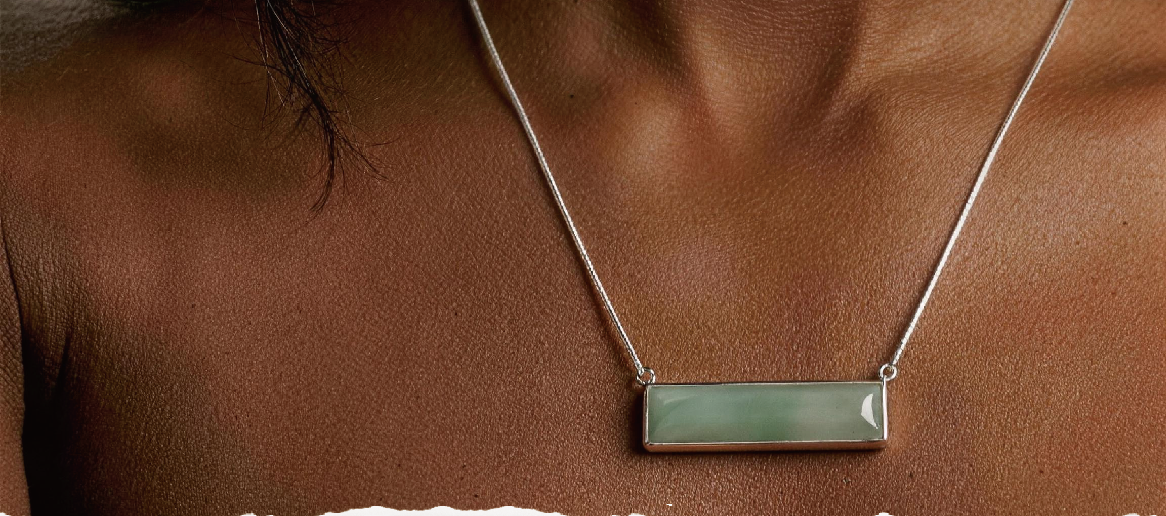 A close-up of a person's chest wearing a silver-toned necklace with a horizontal bar pendant made of pale green stone.